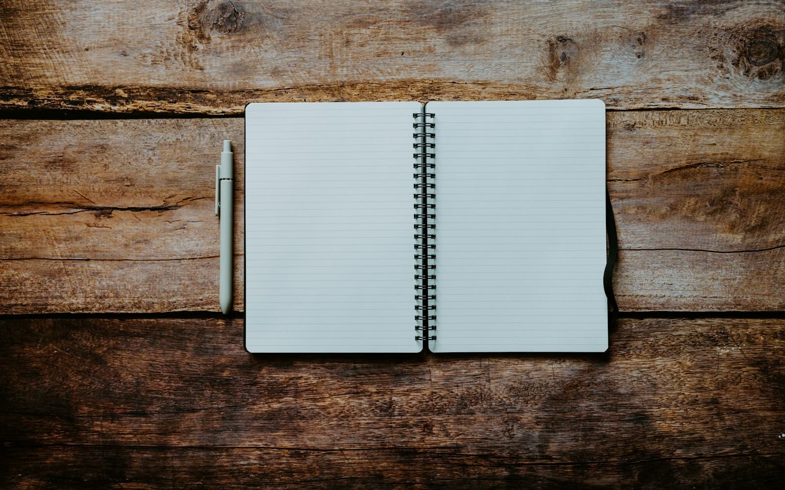 An open spiral notebook and pen resting on a raw wooden table in soft daylight