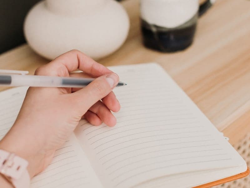 A hand writing notes in a notebook beside a cup of coffee in soft natural light